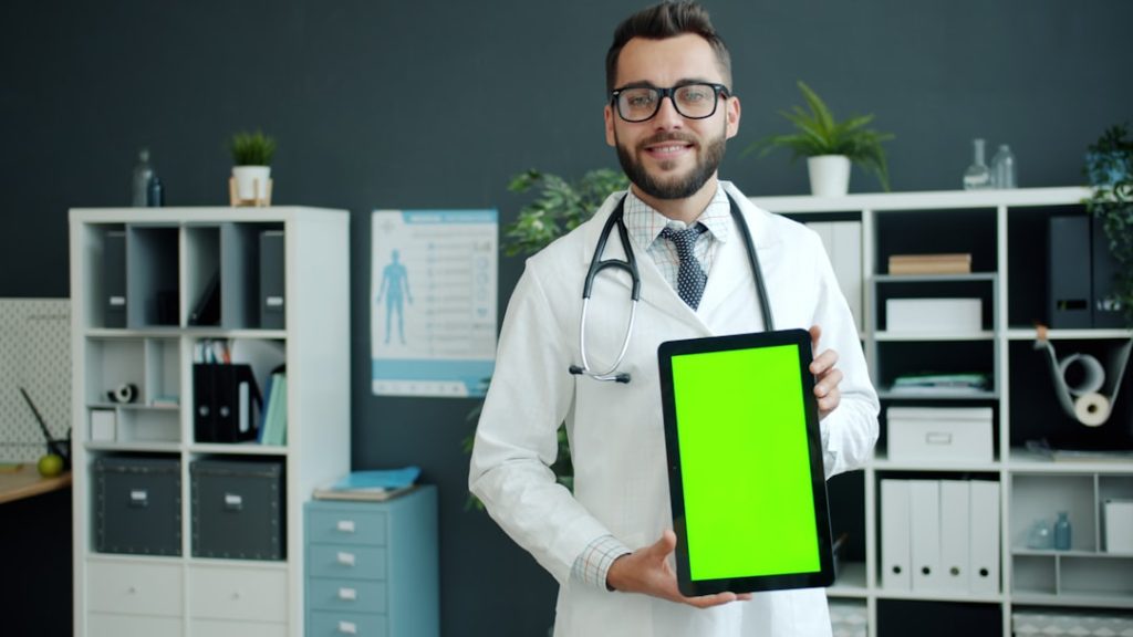 Cheerful young man doctor in white gown is holding tablet with green chroma key screen smiling looking at camera in office in hospital. Gadgets and healthcare concept.
