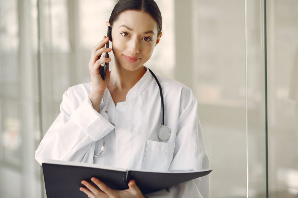 Crop ethnic female doctor in medical uniform with stethoscope and notebook talking on cellphone while standing in clinic corridor