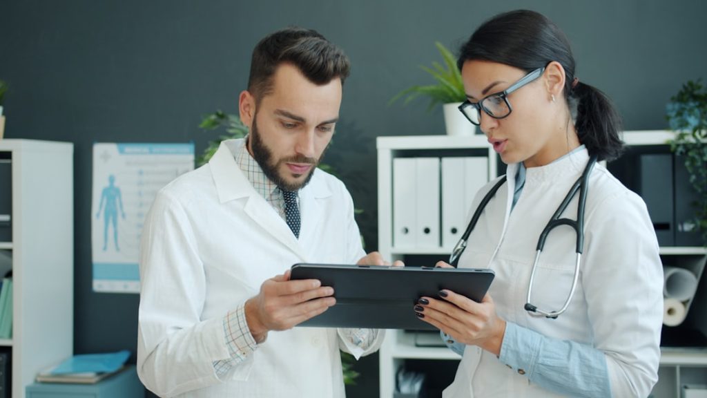 Medics in white gowns young man and woman are using tablet at work talking using gadget in office. Modern devices, people and technology concept.