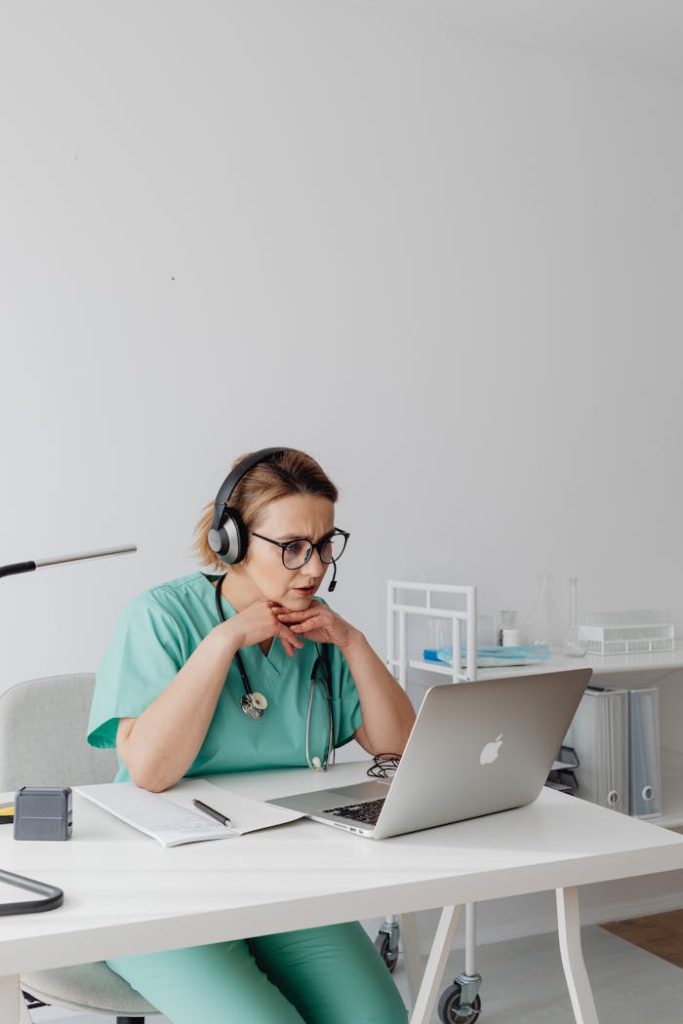 A medical professional focuses intently on a laptop while wearing teal scrubs and headphones.